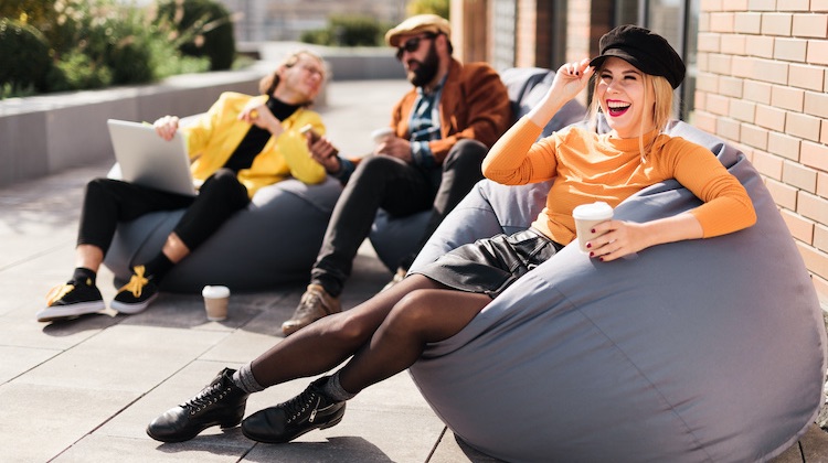 Woman in orange top and leather skirt and boots taps on her black newsboy cap while lounging in a beanbag chair with a coffee cup on a roof deck with two colleagues in the background.