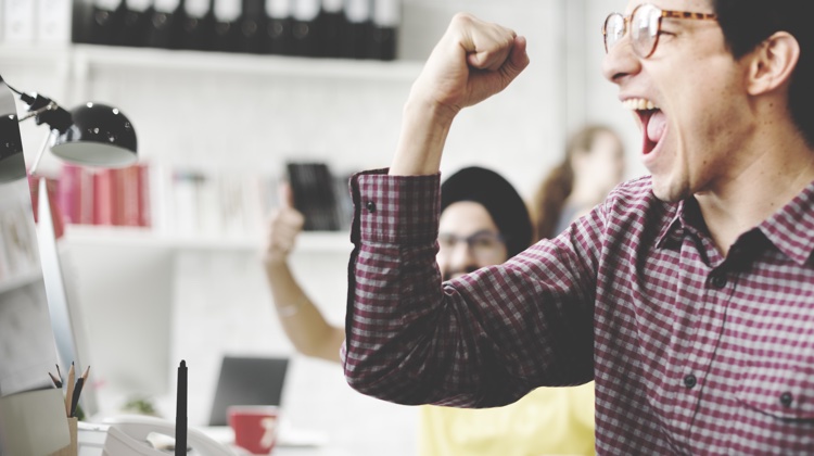 Man in glasses and checkered shirt raises his right fist in celebration while his mouth is open as if he's yelling something in excitement.