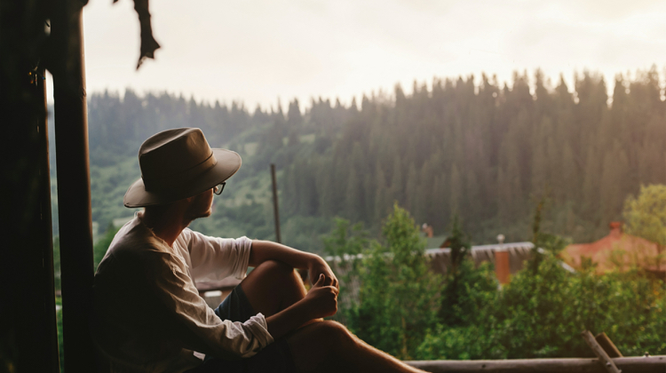 Person leans against wall with a wide-brim hat and stares out at the forest at dusk.