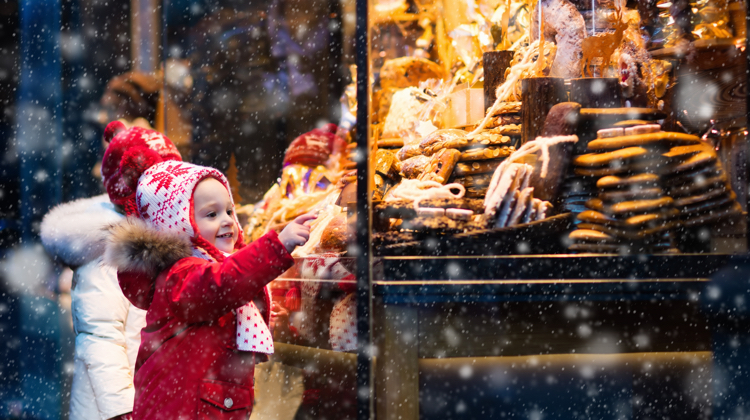 Small child in red snowsuit and hat with tassels reaches for sweets behind a window of a shop as it snows.