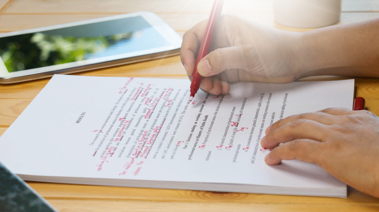 A person is holding a black pen writing on a white legal pad on a desk. In the background there is a tablet sitting on the desk.