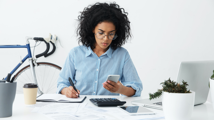 Black woman with curly black hair and wire-rimed glasses looks at cellphone while writing in notebook with tablet, calculator, and other papers on the desk in front of her.