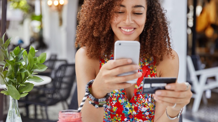 Woman with curly hair smiles as she looks at her phone in her left hand and holds a credit card in her right hand.