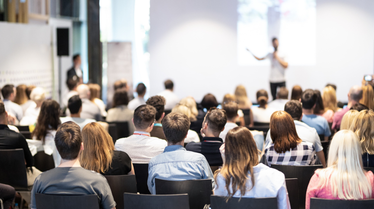 A group of people sits in an conference space facing a speaker at the front of the room.