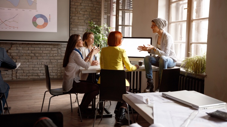 Three women sit in an open off around a table while a fourth sits on a window sill explaining something to the group.