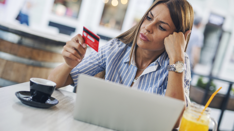 A woman is sitting in a coffee shop at a table looking at her credit card. On her table there is a cup of coffee, a laptop, and an orange drink in a mason jar.