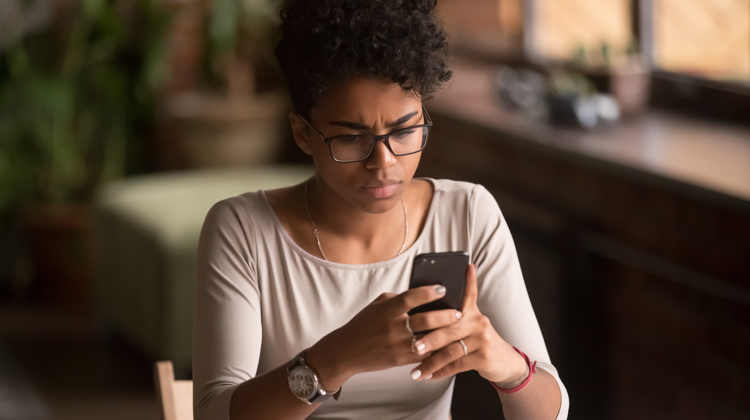 A woman wearing glasses, a silver necklace, a pink shirt, a red bracelet, and a wrist watch is sitting down staring at a smartphone. The background behind her is blurry.