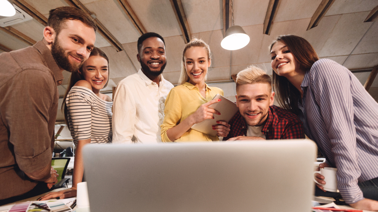 A group of men and women are surrounding a laptop. They are all looking at the screen smiling. They are at a desk in an office space.