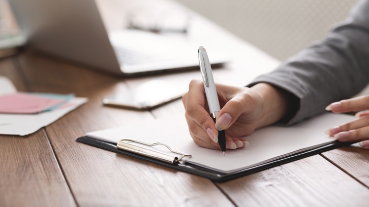 Someone is writing with a black and white pen on a piece of paper attached to a clipboard. The person has a gray suit on and there is a silver laptop and some scattered documents in the background.