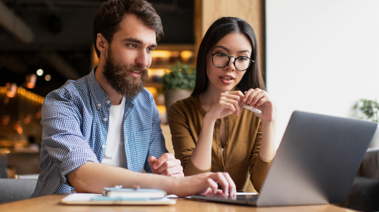 A man and woman looking at an open laptop. Behind them, is a coffee shop with ambient lighting.