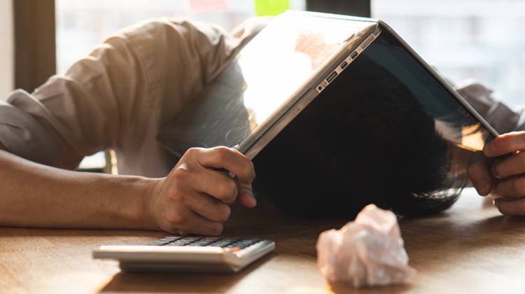 A person with their face down on a desk holding their laptop over their head. In front of them there is a calculator and a crumpled up piece of paper.