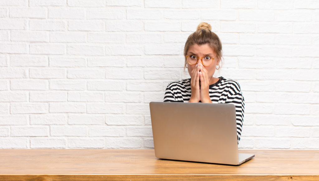 Blonde woman with hair tied in bun in black-and-white striped top and mustard-rimmed glasses covers her mouth with her hands in shock while a laptop sits open in front of her.