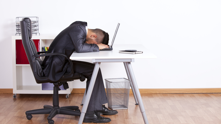 Man in suit sits in a rolling chair at a metal desk with his head laying face down on his laptop.