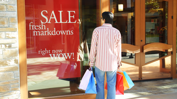 Man in red and white vertical striped shirt and jeans holds five shopping bags of various colors while standing in front of store window that says "sale fresh markdowns."