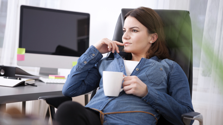 A woman is sitting in an office chair at a desk with a computer on it. She is wearing a jean jacket and holding a white coffee mug.