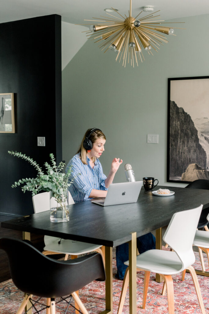 Nicki, one of the hosts of the Build Your Copywriting Business podcast, sits at a black table with six chairs, four white, two black, as she records a podcast episode with a microphone, laptop, and doughnut in front of her.