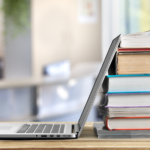 Laptop on a desk leaning against a stack of 8 books of different shapes and sizes.