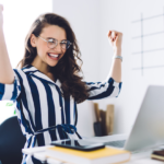 Woman in blue and white striped dress sitting in front of her laptop has arms up in celebration