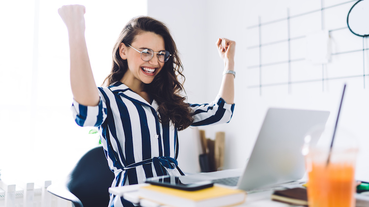 Woman in blue and white striped dress sitting in front of her laptop has arms up in celebration.