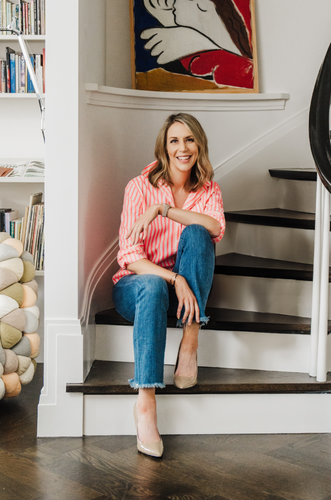 Nicki Krawczyk, founder of the Comprehensive Copywriting Academy, sits on a staircase in jeans and a striped coral button-down top.