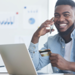 Man on cell phone with credit card in hand sits in front of his laptop