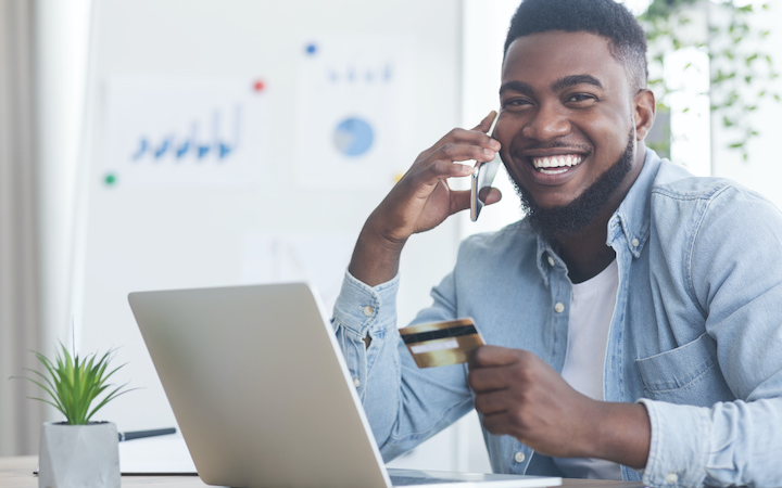 Man on cell phone with credit card in hand sits in front of his laptop