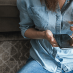 Image is cropped showing a person's torso wearing a denim top and jeans as they sit on a carpet holding a phone and pointing a finger at the screen.