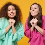 Two women against mustard backdrop, one in green shirt one in pink, hold hands together in pleading motion.