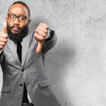 Man in grey suit against concrete wall has one thumb up and one thumb down.