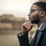 Man in glasses wearing a suite with his hand to his chin stares off reflectively in the distance