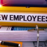 Yellow binder that says "new employees" sits on a desk on top of other binders and papers.