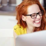 Red-headed woman in black glasses and a yellow shirt laughs in front of an open laptop