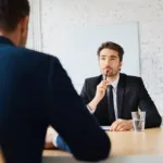 Two men in suits sit across a table facing each other with one holding a pen to his mouth pensively and a glass of water sits on the table