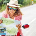 A woman on a moped, wearing a hat and glasses, leans on the handlebars and looks at a map.