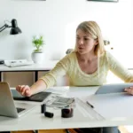 Woman sits at desk with a pen in her left hand and right hand hovering over her laptop.