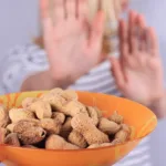 A hand in the foreground holds an orange bowl filled with peanuts while a person in the background holds up two hands blocking the bowl from them.