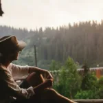 Person leans against wall with a wide-brim hat and stares out at the forest at dusk.