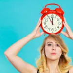 Blond woman holding an old-fashioned red clock on top of her head