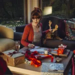 Woman sits in front of a couch, where a dog sits with reindeer antlers, writing on a piece of paper while the coffee table in front of her is strewn with ribbon, gifts, and a glass of sparkling wine.