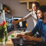 Woman in blazer leans over a designer's desk pointing at one of the two monitors while the designer uses a stylus to make an update.