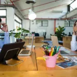 A frustrated-looking woman with her head in her hands faces a man across a desk who is scratching his head