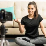 A woman is sitting cross-legged on the floor in front of a cream colored couch. She is looking at a camera mounted on a tripod. The woman is wearing a black shirt and gray leggings.