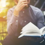 A man is sitting outdoors and the sun is beginning to set behind him. He is wearing a blue button up shirt and reading a book.