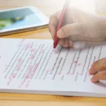 A person is holding a black pen writing on a white legal pad on a desk. In the background there is a tablet sitting on the desk.