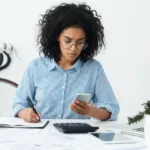 Black woman with curly black hair and wire-rimed glasses looks at cellphone while writing in notebook with tablet, calculator, and other papers on the desk in front of her.