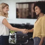 Two women in their thirties shaking hands and smiling in a modern conference room