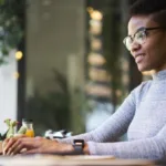 Woman in glasses and a grey turtleneck sits at a desk typing.