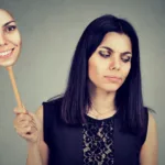 Woman holds up a stick with a copy of her face smiling on it as she looks down to the other side frowning