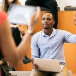 Woman stands at front of a classroom with tiered seating while focus of image is on a man raising his hand with a laptop resting on his lap.