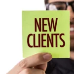 Man with glasses and beard holds a lime green sticky note in front of his face that says "new clients" in all capital block letters.